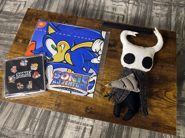 An image of a handmade Hollow Knight plushie and the classic game Sonic Adventure on the Dreamcast.